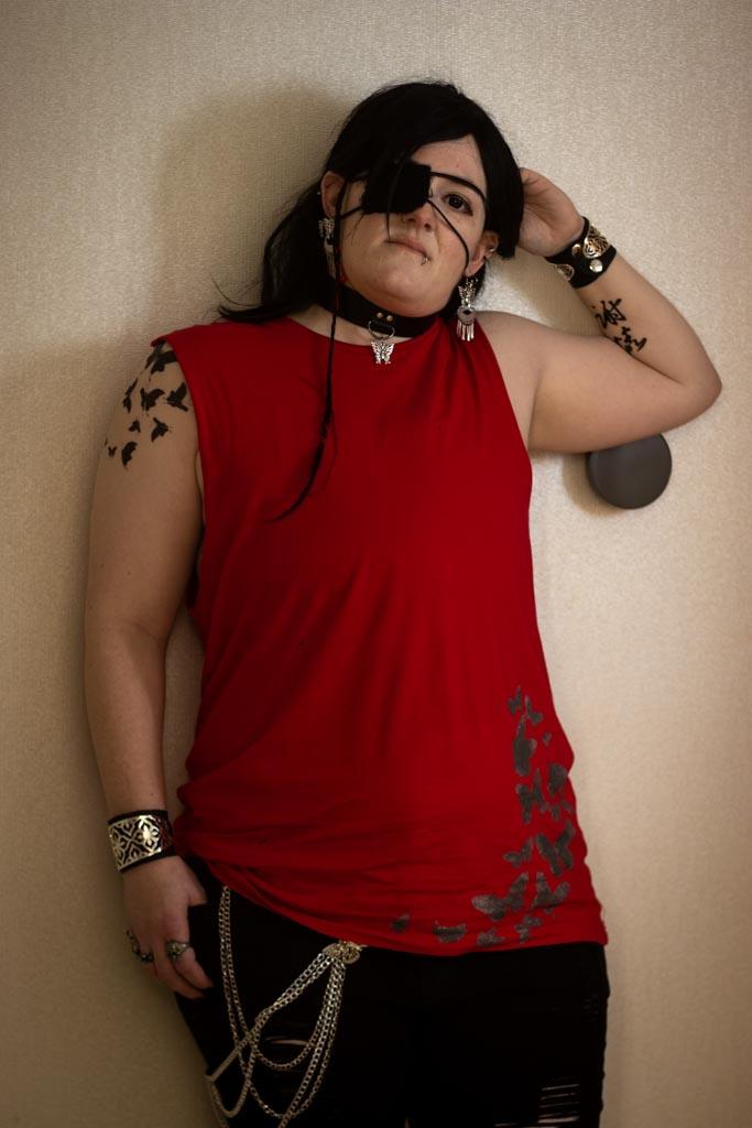 Waist-up shot of modern 'street fashion' Hua Cheng cosplay against an off-white wall, one hand behind the head so a tattoo in Chinese calligraphy is visible on the forearm. 
