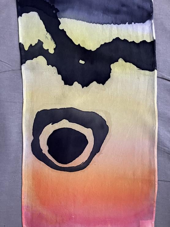 A portion of a silk scarf lays on my ironing board. It is painted in a gradient from red to yellow. On top of that gradient are black shapes. In the middle is a circle surrounded by a ring, and above is a thick jagged black line running across the scarf. 